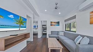 A modern living room with a large TV, a grey sofa, a wooden coffee table, and a view of the beach through a large window. - Discovery Parks - Eden (Boydtown)