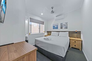 A hotel room with a large bed, a wooden desk, a nightstand with a lamp, and a window with a view of the outside. - Discovery Parks - Eden (Boydtown)