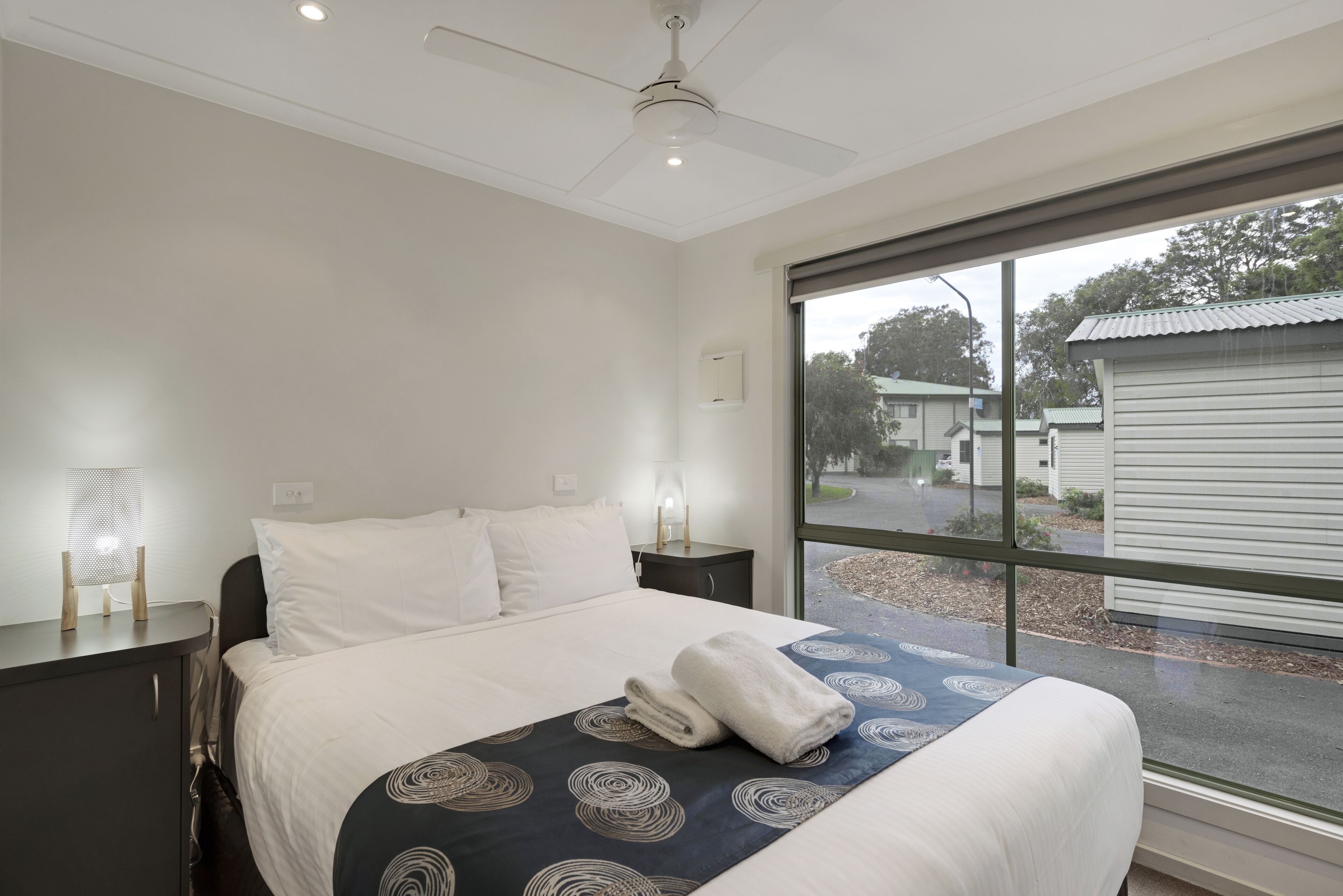 A hotel room with a bed, bedside tables, a ceiling fan, and a large window with a view of the outside.