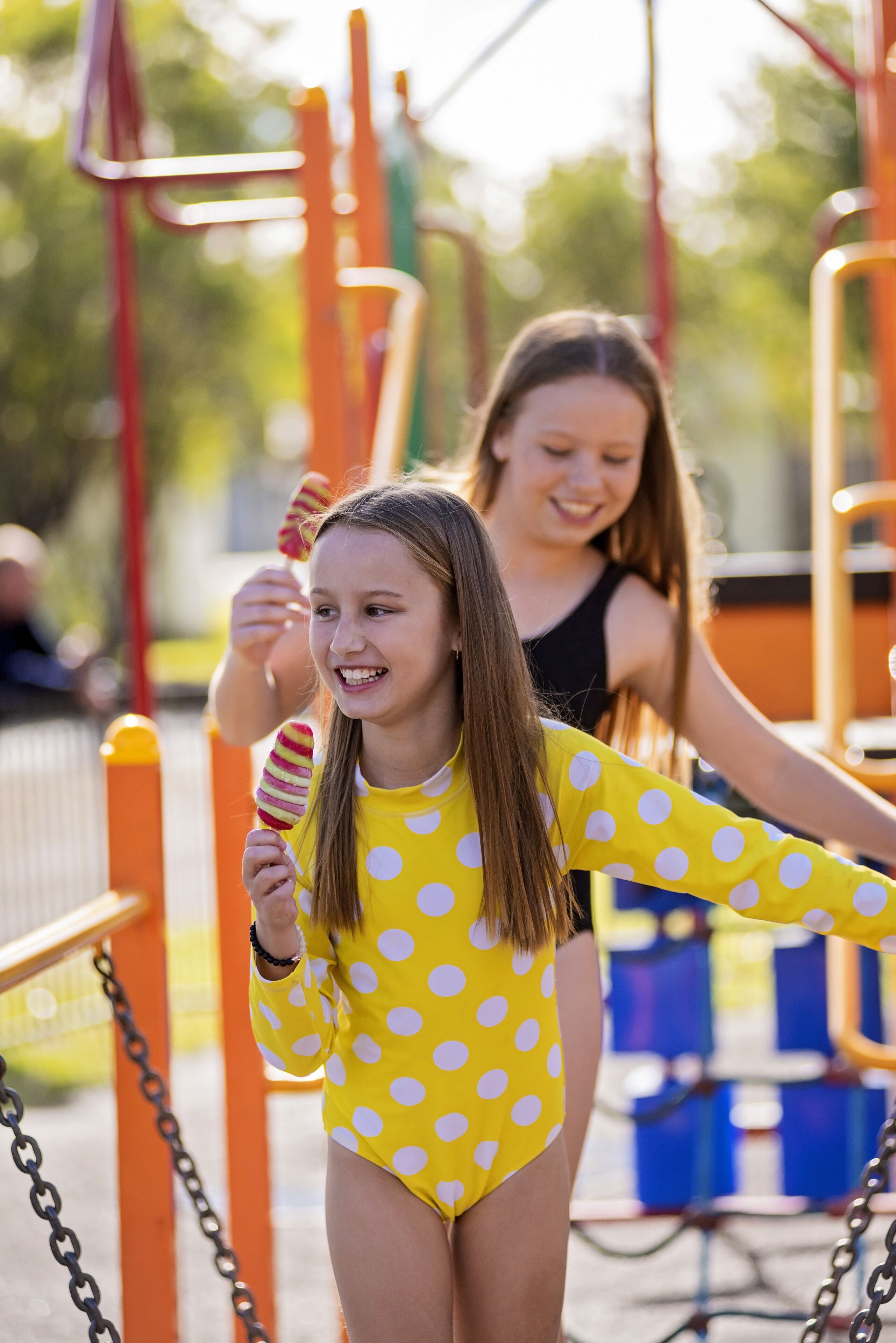 A child in a polka dot swimsuit holding a lollipop at a playground with orange and yellow equipment.