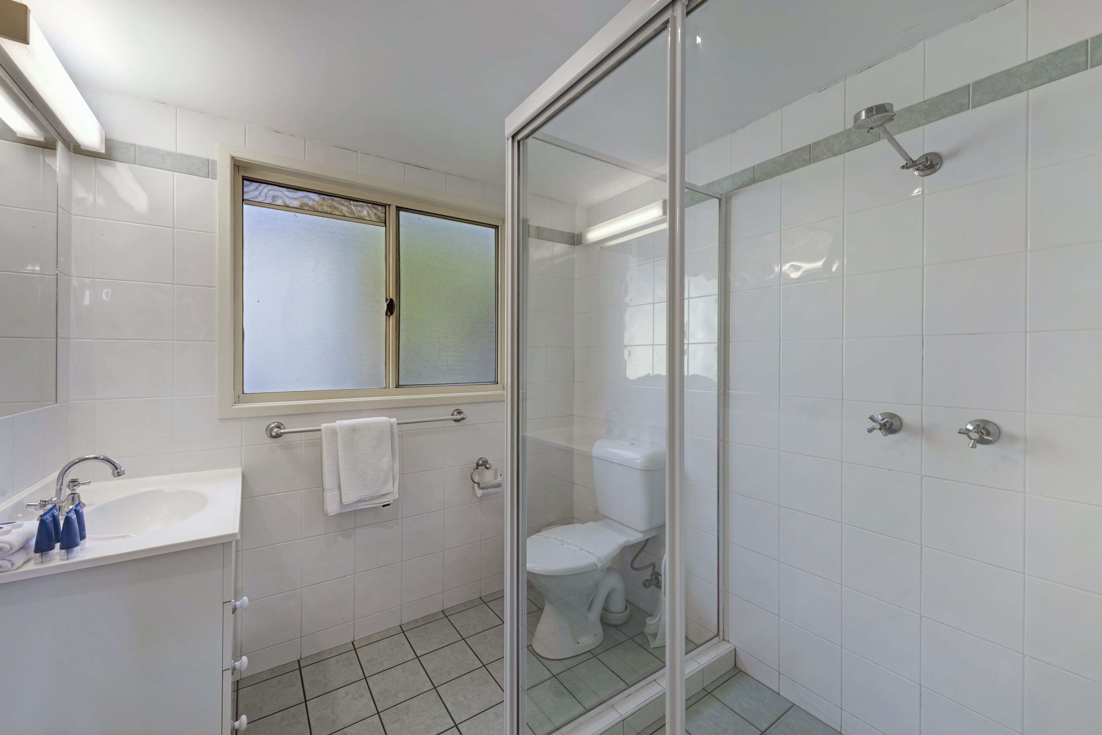 A bathroom with a glass-enclosed shower, a toilet, a sink with blue soap dispensers, and a window above the sink.