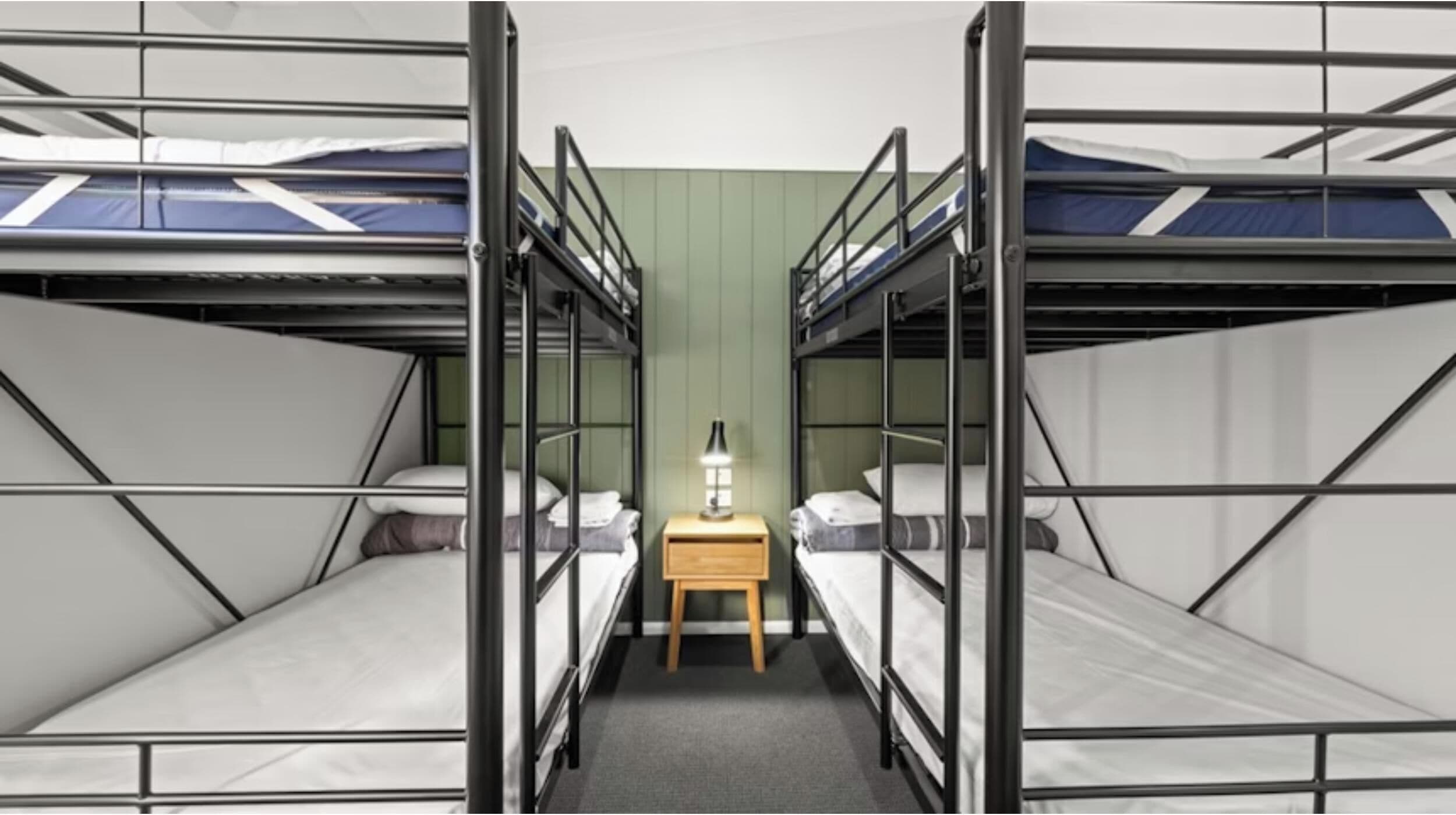 A room with bunk beds arranged in a V-shape, a small wooden nightstand with a lamp, and a green wall.