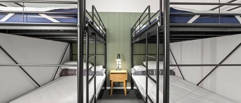 A room with bunk beds arranged in a V-shape, a small wooden nightstand with a lamp, and a green wall.