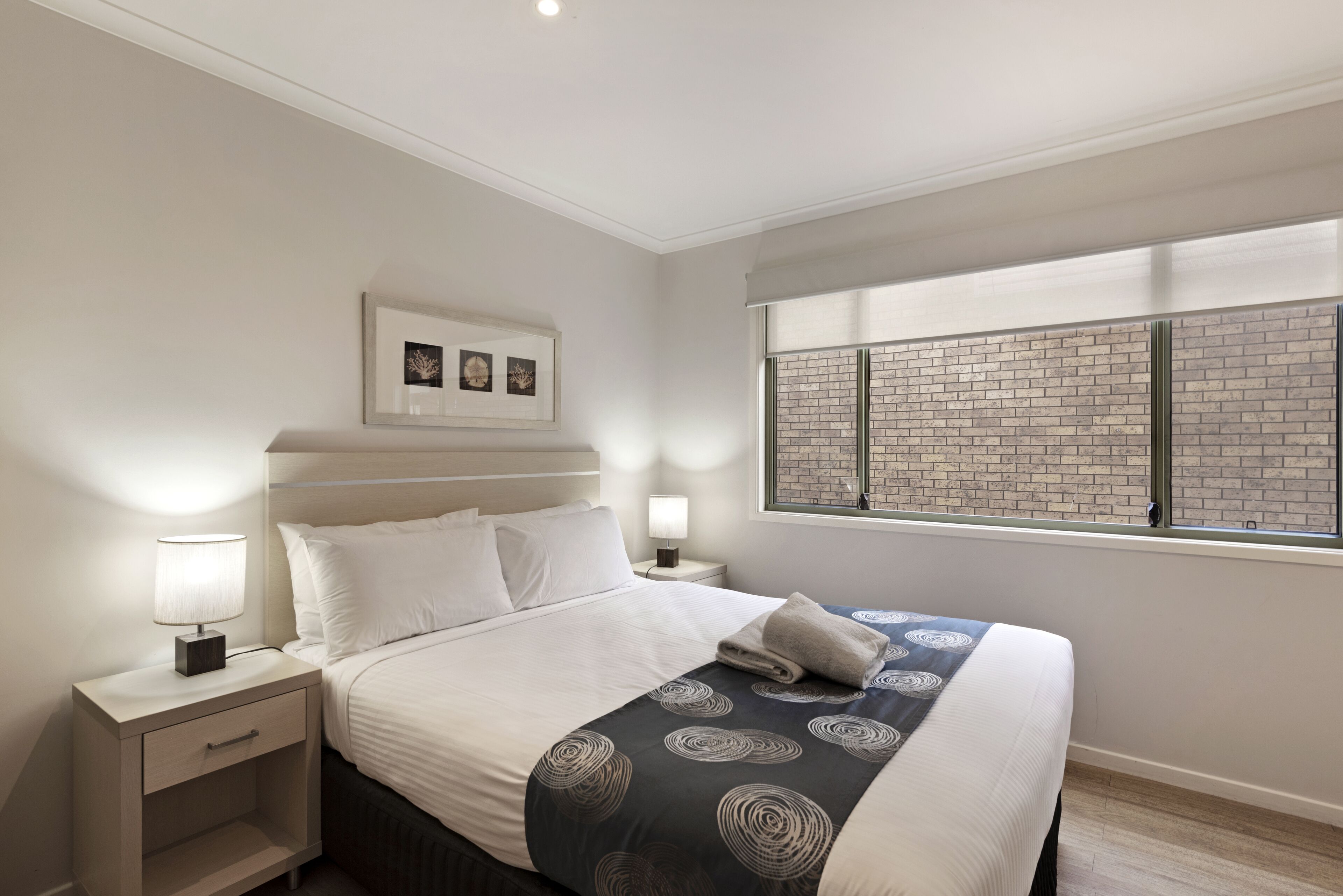 A neatly made bed with a patterned bedspread, a bedside table with a lamp, and a window with a brick wall view.