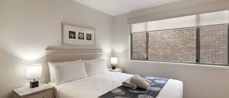 A neatly made bed with a patterned bedspread, a bedside table with a lamp, and a window with a brick wall view.