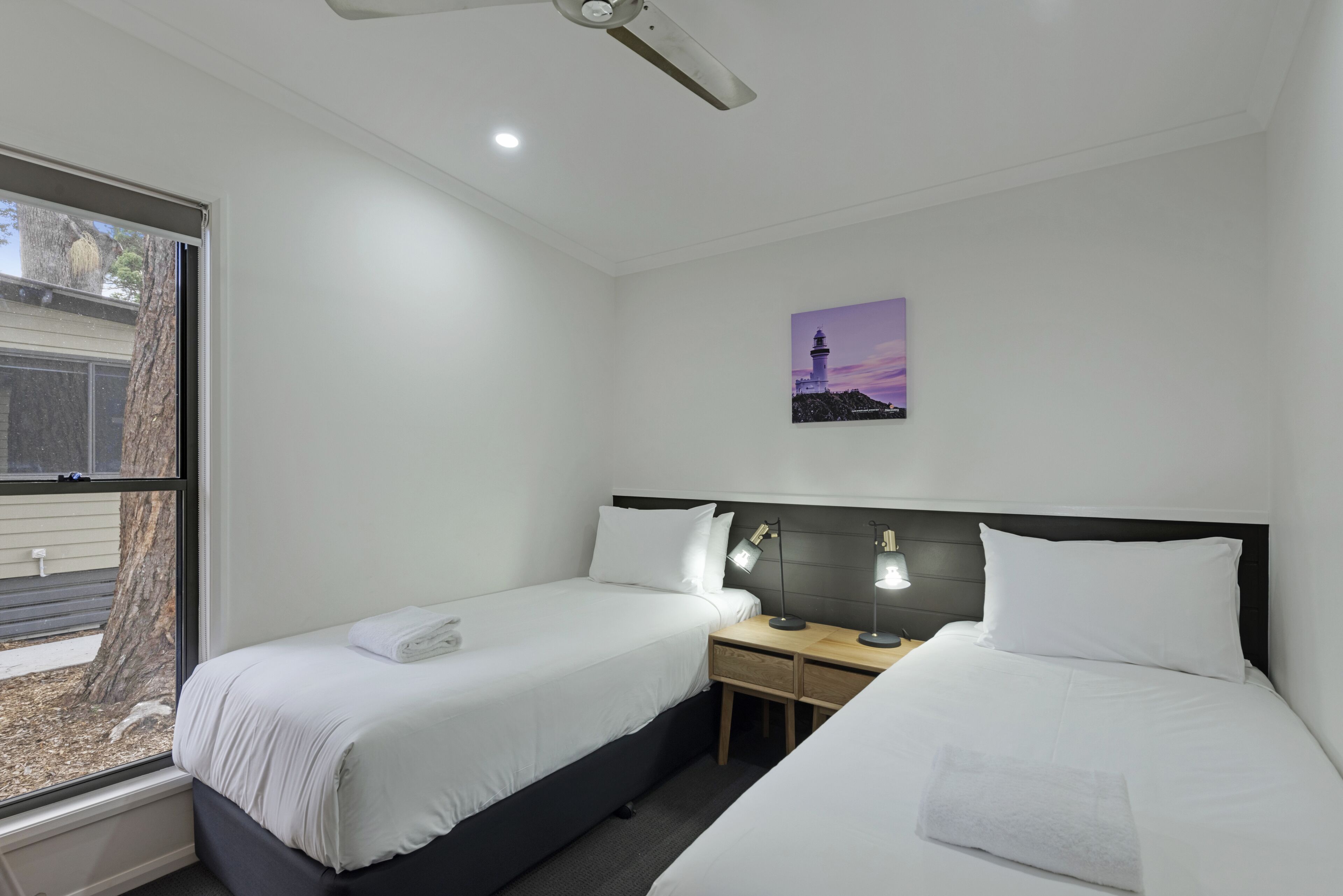 Quarto de hotel com duas camas de solteiro, uma mesa de cabeceira com abajur, uma janela com vista para uma árvore e um edifício, e um ventilador de teto.