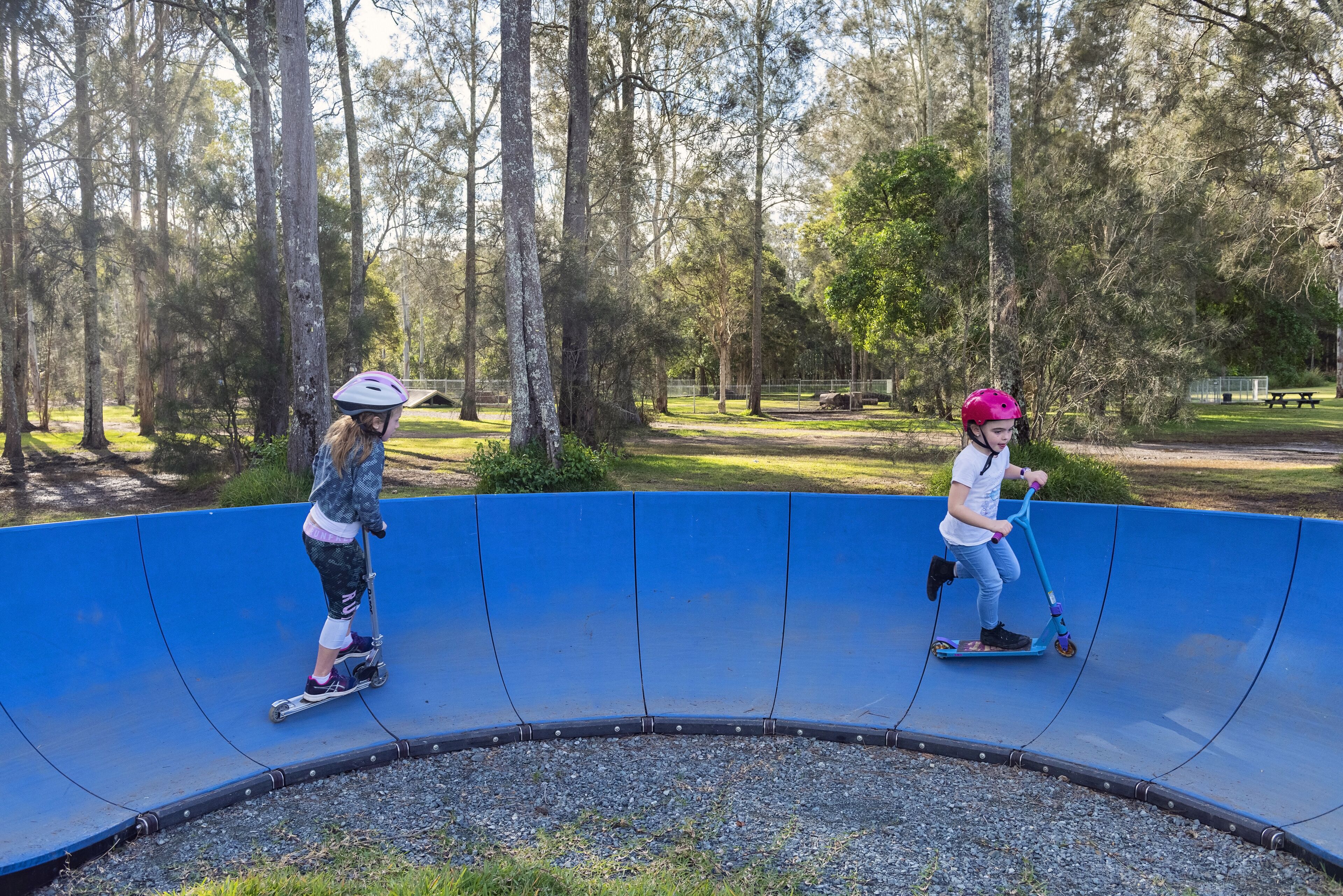 Two children riding scooters on a blue track in a park with trees and a picnic area in the background.