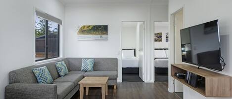 Un salon moderne avec un canapé d’angle gris, une table basse en bois, un téléviseur à écran plat posé sur une étagère en bois et une grande fenêtre.