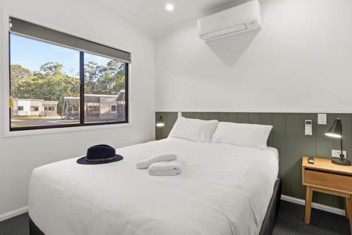Habitación de hotel con cama, mesita de noche, lámpara, ventana con vistas a edificios y unidad de aire acondicionado.