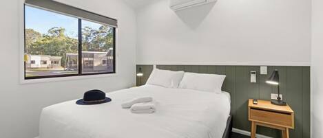 A hotel room with a bed, a nightstand, a lamp, a window with a view of buildings, and an air conditioning unit.