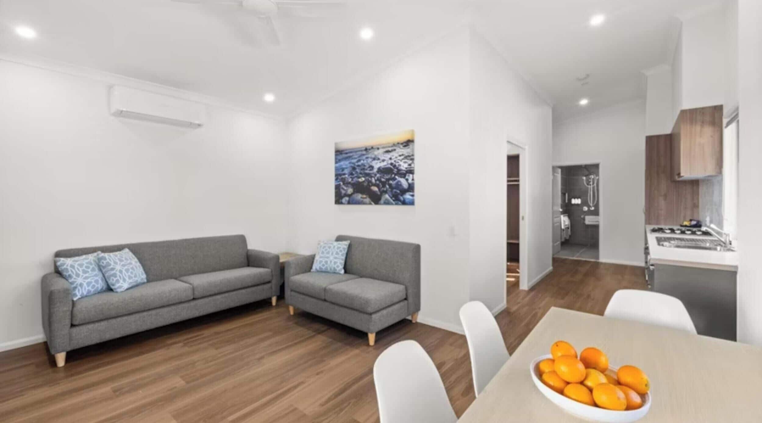 Un salon moderne avec un canapé gris, une table à manger sur laquelle se trouve un saladier rempli d’oranges, et un coin cuisine visible à travers une porte ouverte.
