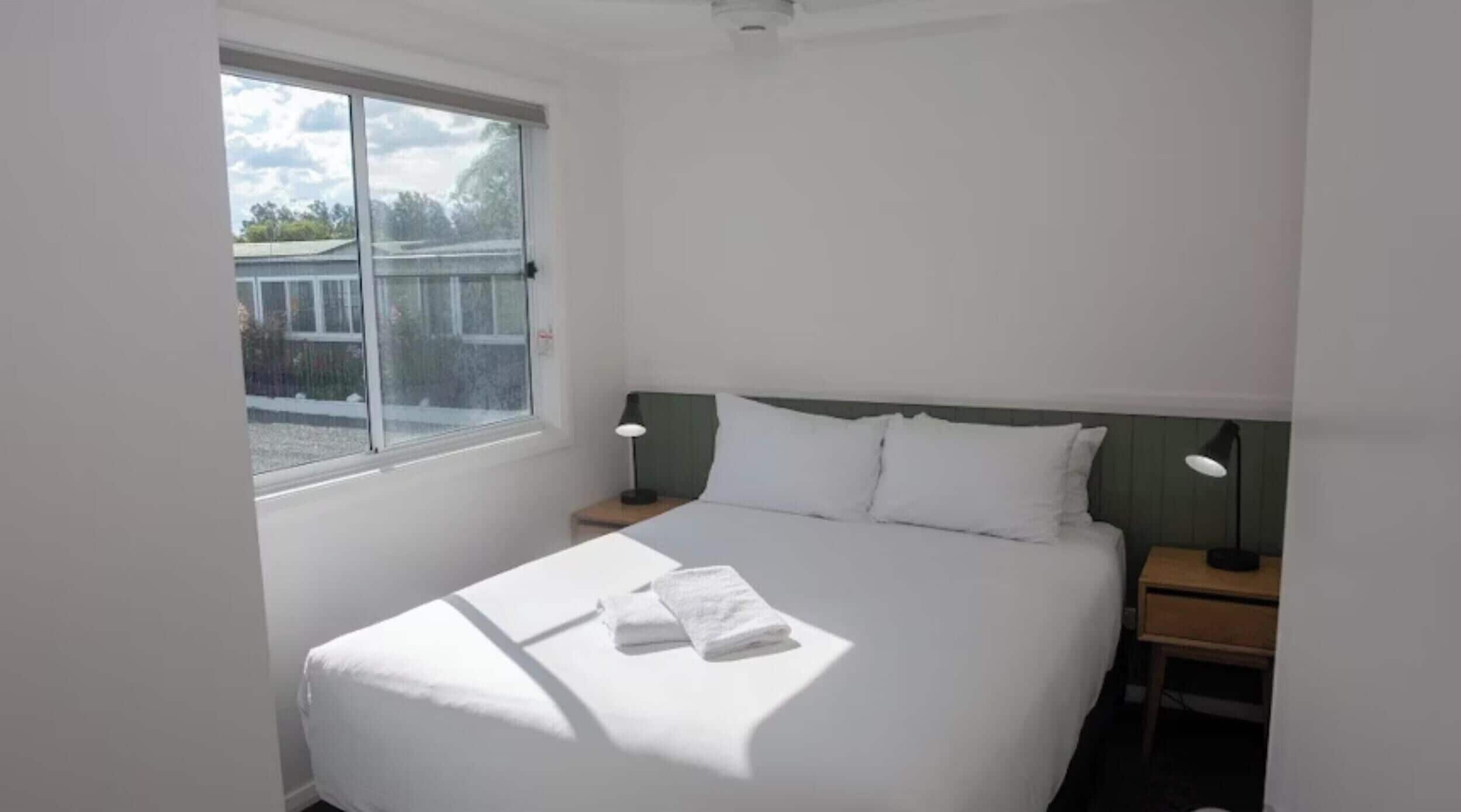 Una habitación de hotel con una cama grande, dos lámparas de noche, un ventanal con vistas a edificios y una lámpara de techo.