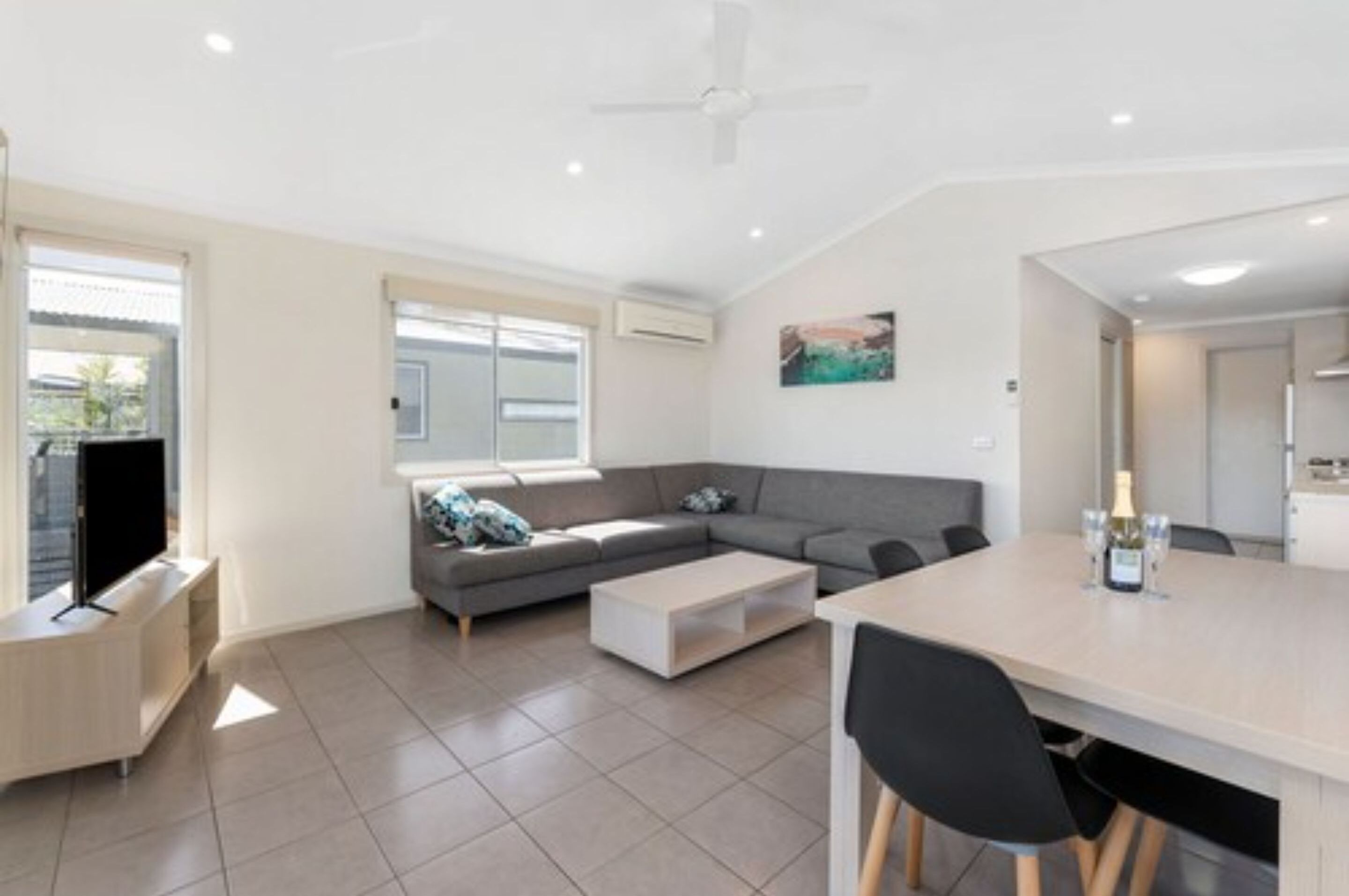 A modern living room with a grey sectional sofa, a flat-screen TV, a coffee table, and a dining area with a table and chairs.