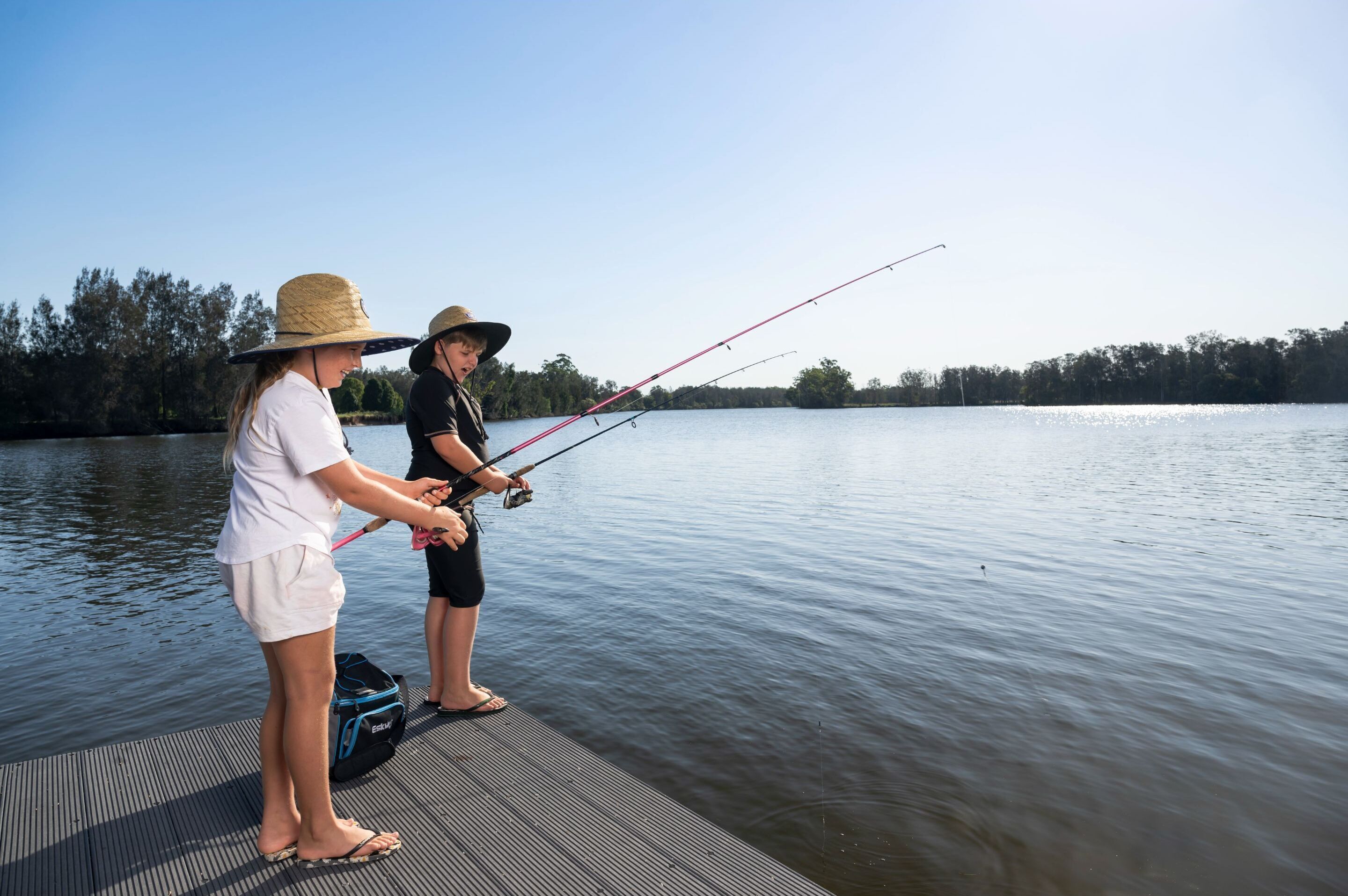 Two people fishing on a dock by a calm lake with trees in the background.