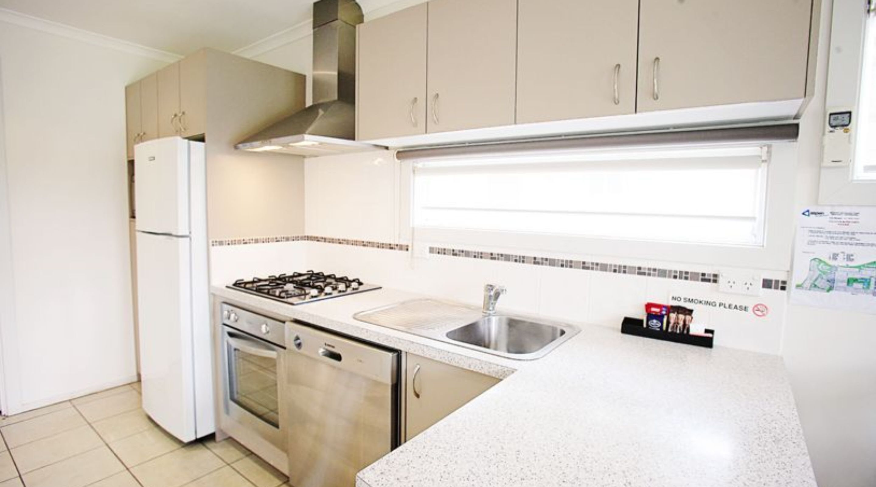 A compact kitchen with a gas stove, oven, and sink. There is a stainless steel refrigerator and cabinets with handles. A 'No Smoking' sign is visible on the wall.