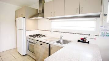 A compact kitchen with a gas stove, oven, and sink. There is a stainless steel refrigerator and cabinets with handles. A 'No Smoking' sign is visible on the wall.
