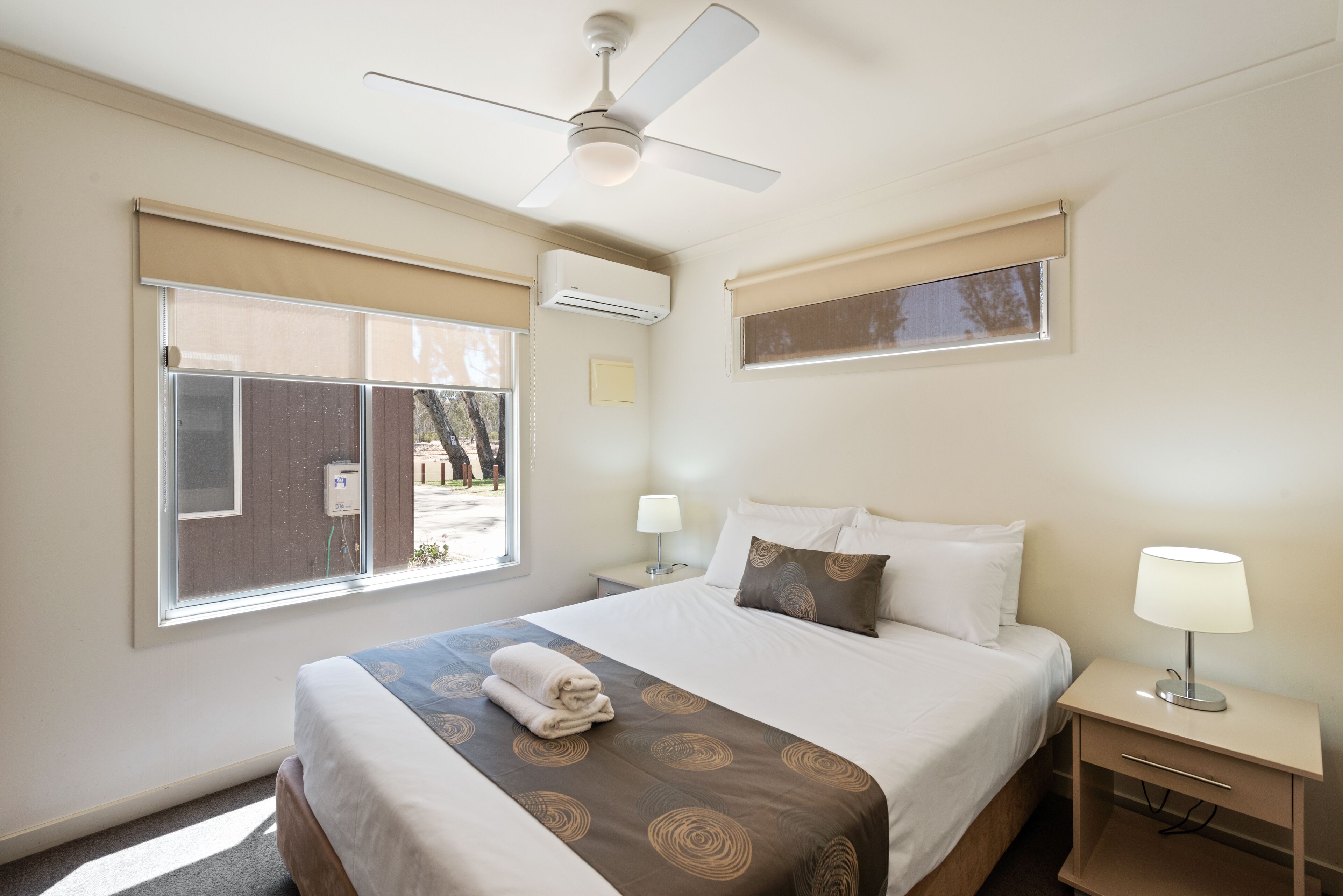 A hotel room with a bed, a nightstand, a lamp, an air conditioning unit, a ceiling fan, and a window with blinds.