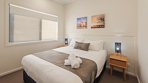 A neatly made bed with a brown and white bedspread, two wall-mounted paintings, and a bedside table with a lamp.