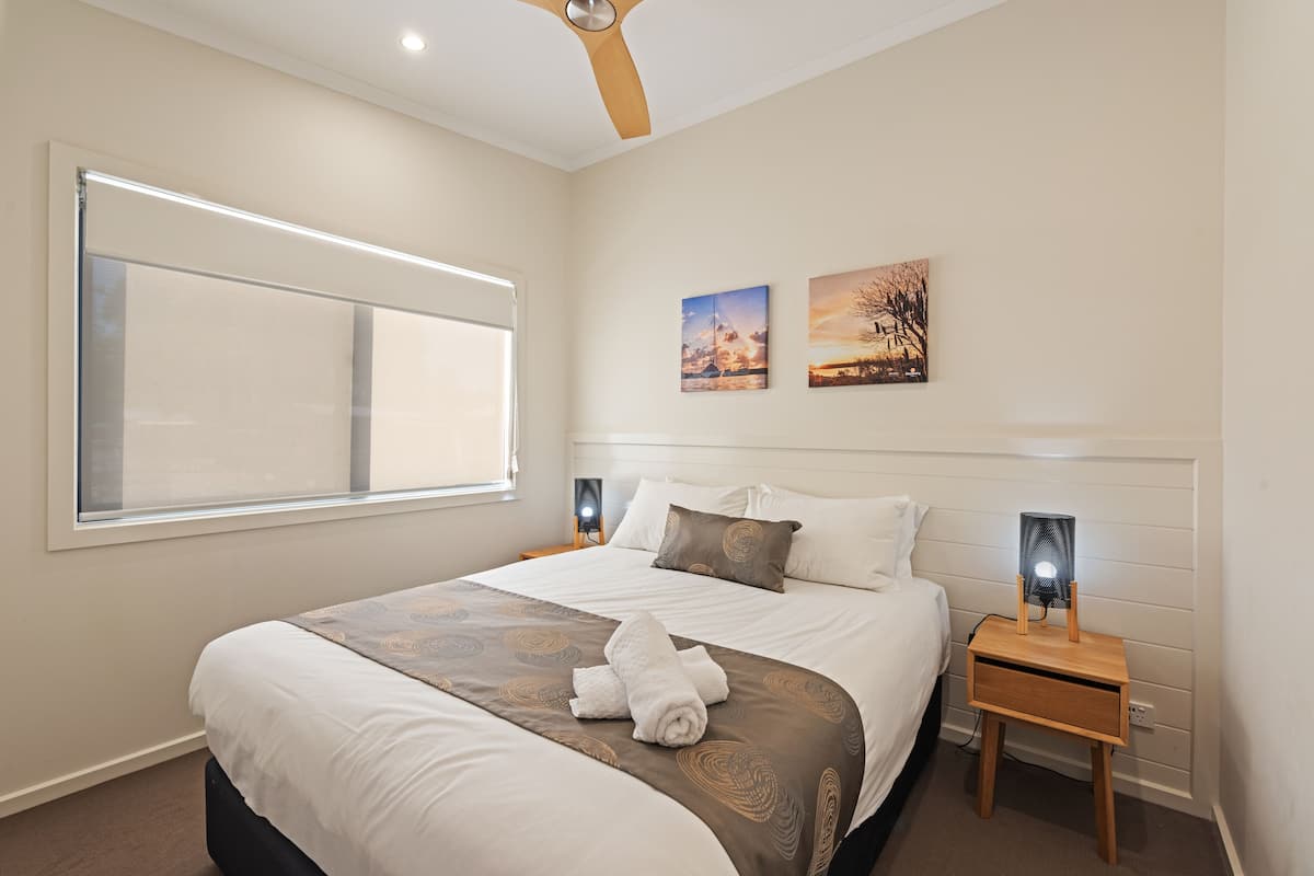 A neatly made bed with a brown and white bedspread, two wall-mounted paintings, and a bedside table with a lamp.