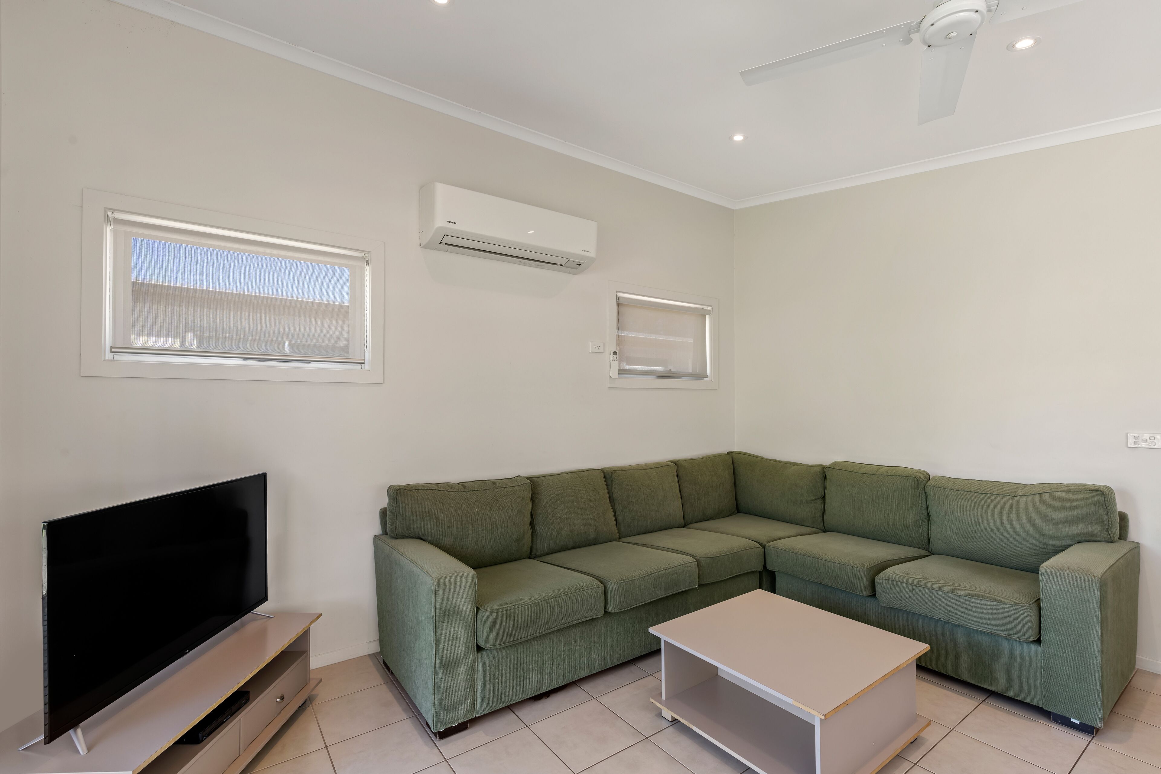 A living room with a green sofa, a flat-screen TV, a ceiling fan, and two windows.