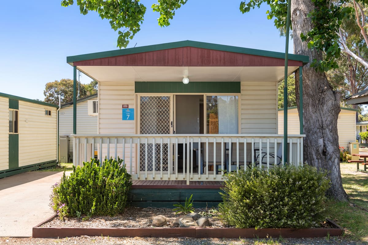 A single-story mobile home with a porch, a small garden with rocks and shrubs, and a tree to the right.