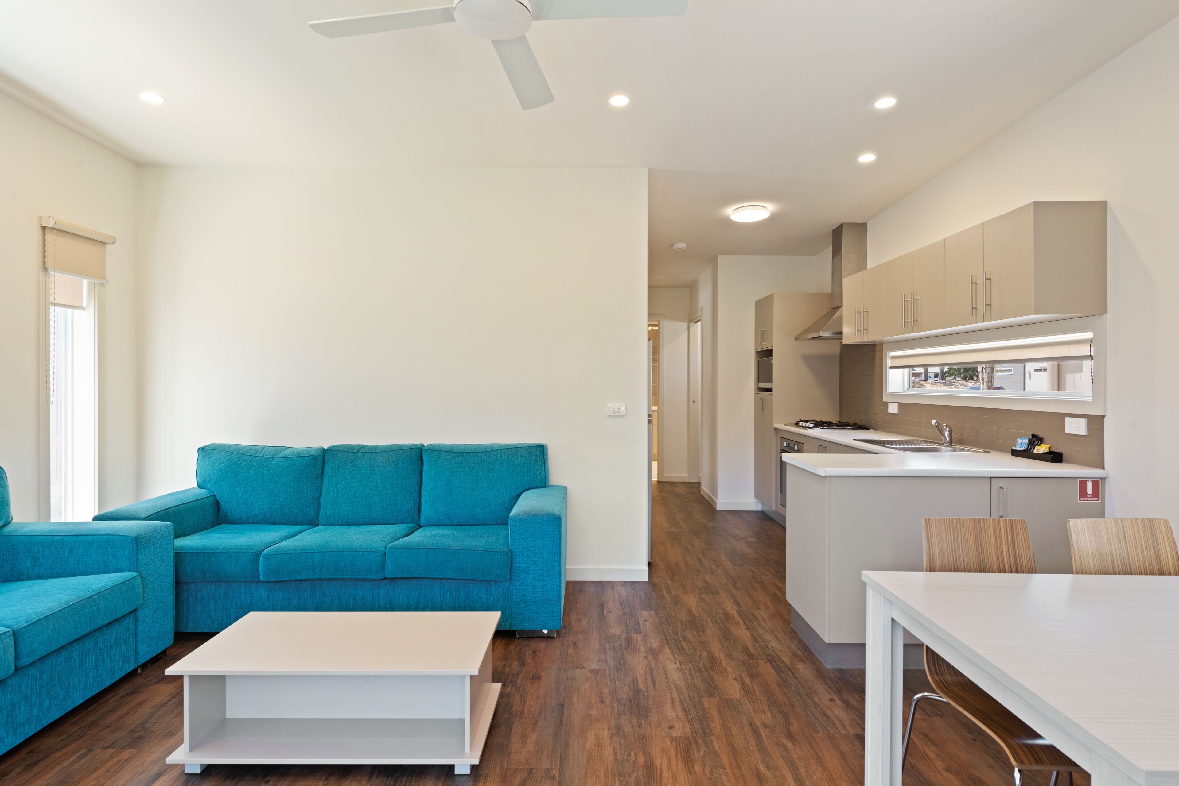 A modern living room with a teal sofa, a white coffee table, and a kitchen area with wooden cabinets and a white countertop.