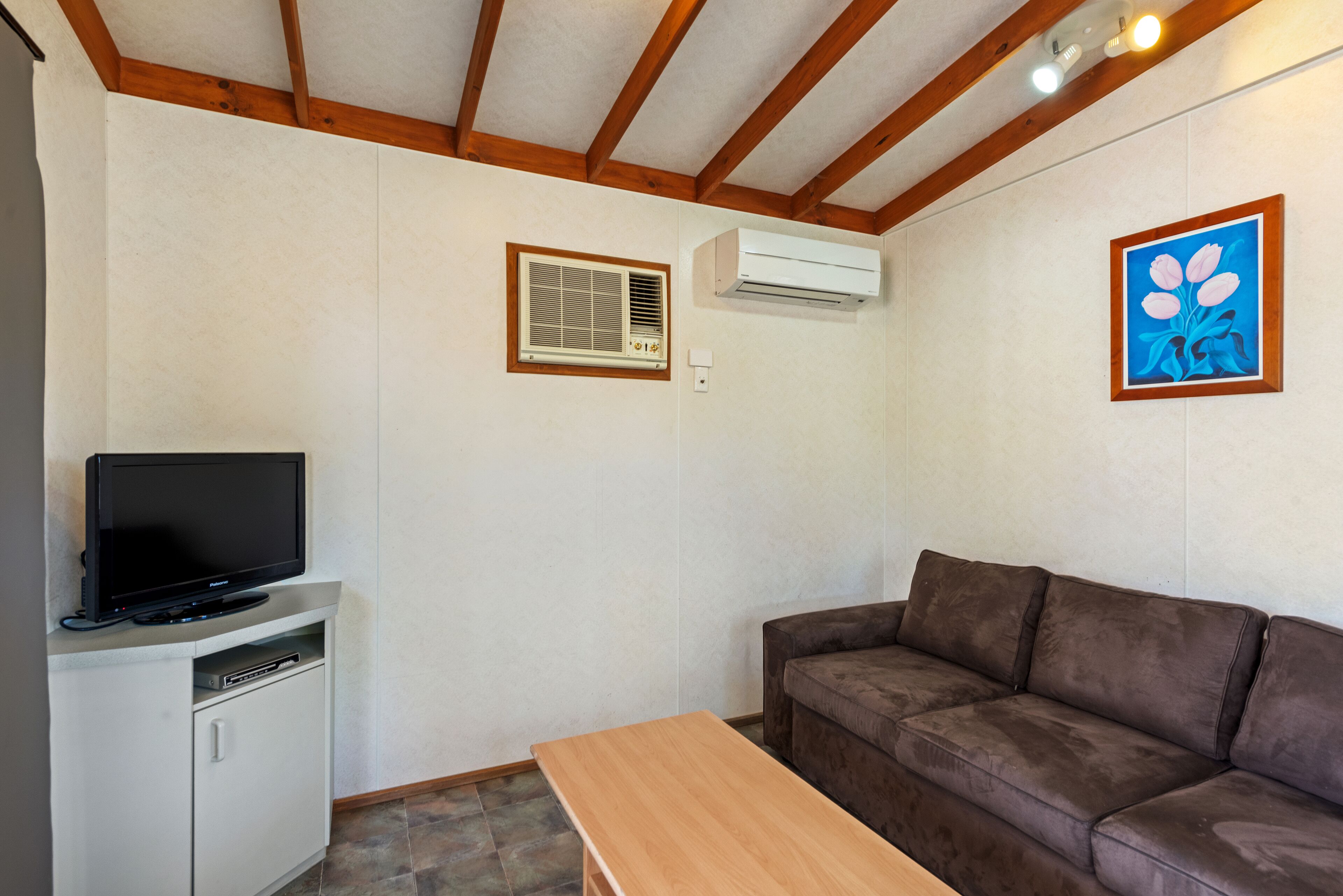 A room with a brown sofa, a wooden table, a TV on a cabinet, an air conditioner, and a painting on the wall.