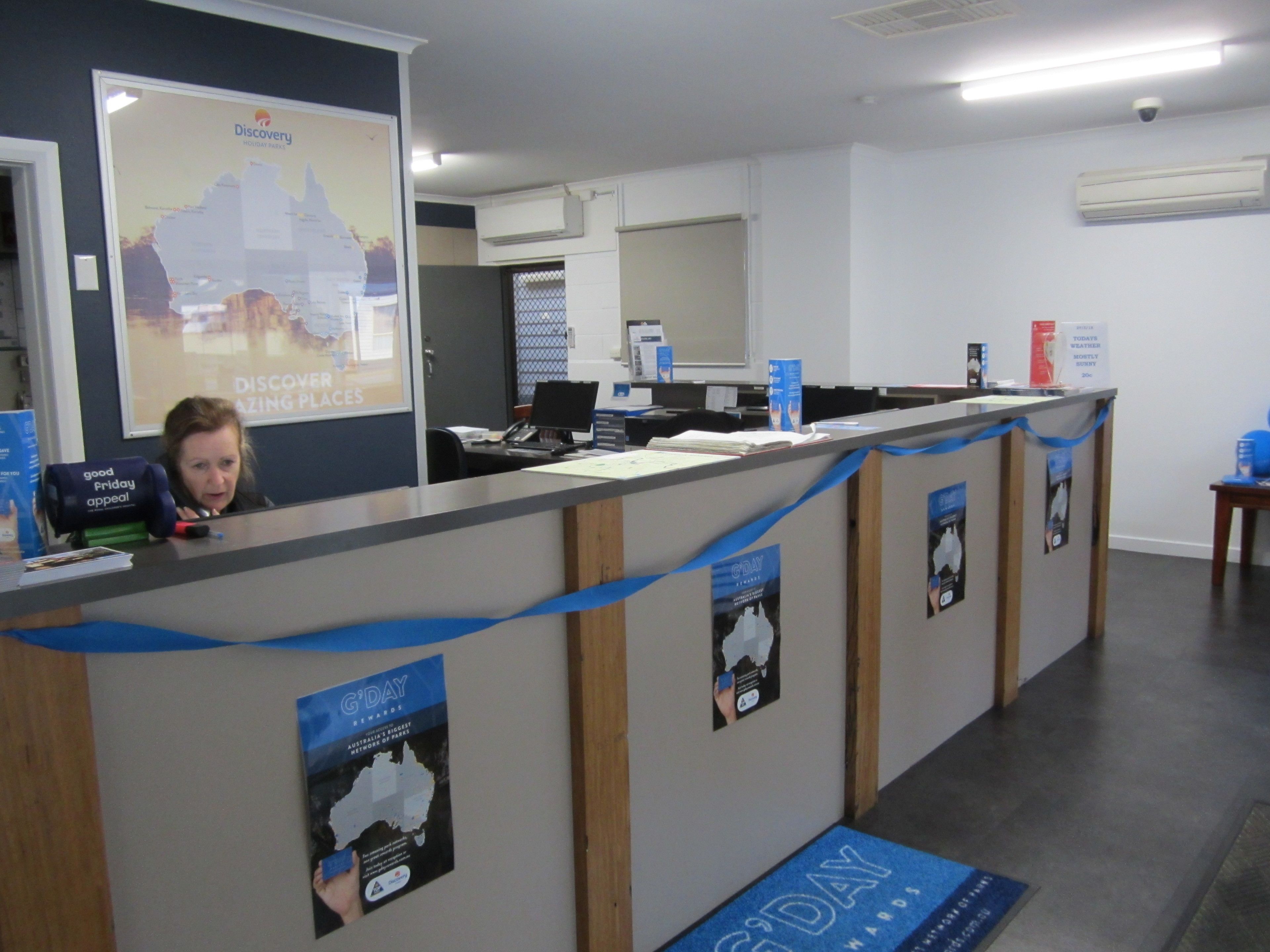 A reception desk with a map of Australia, a computer, and a poster for G'DAY Rewards.