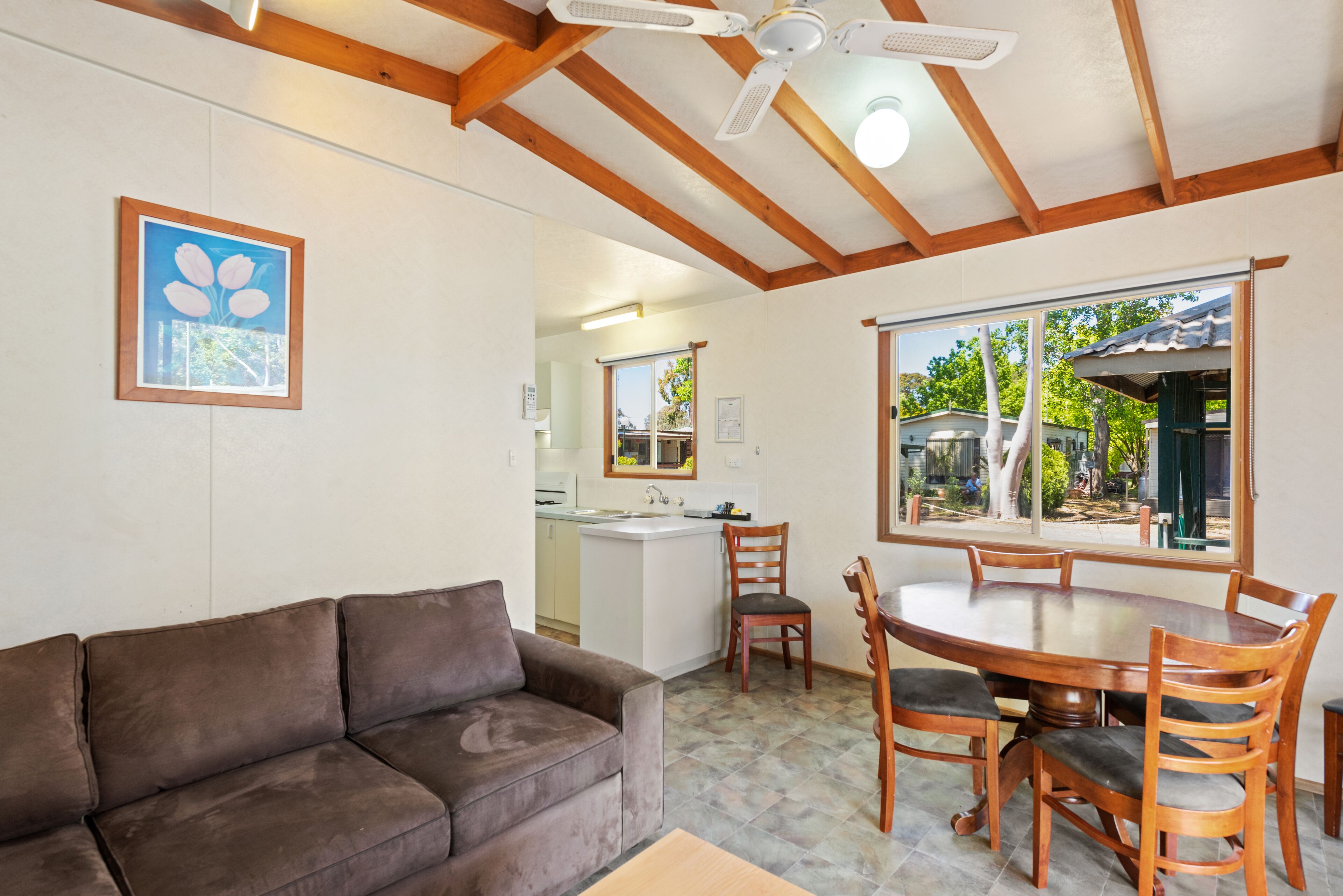 A cozy living room with a brown sofa, wooden dining table with chairs, a kitchen area, and a large window with a view of trees.
