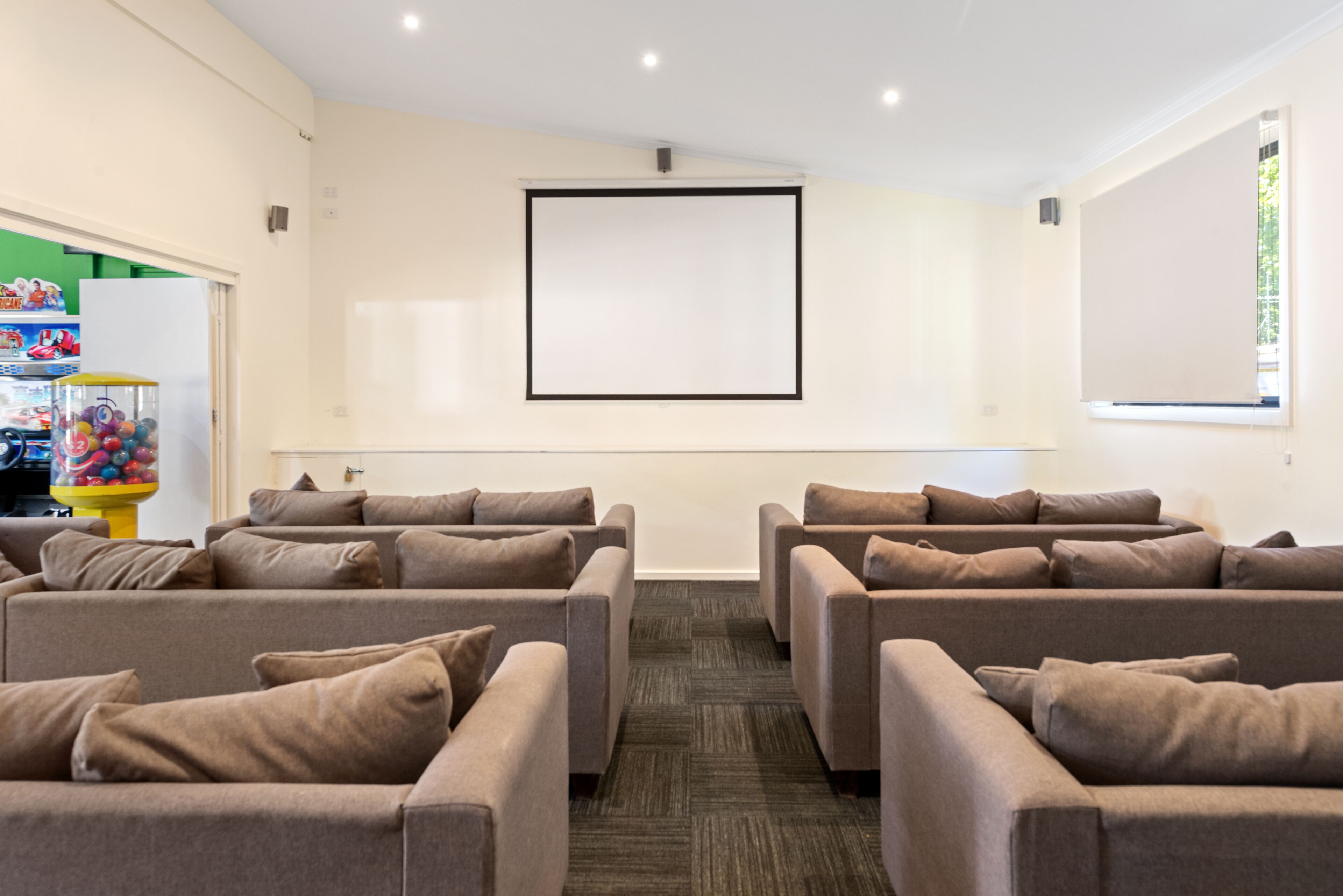 A room with multiple sofas arranged in a semi-circle, a large blank screen on the wall, and a vending machine with colorful items on the left.