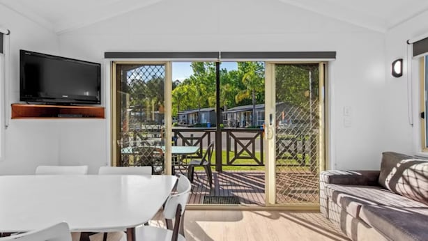 A modern room with a white table, a sofa, a TV mounted on the wall, and a sliding glass door leading to an outdoor area with a view of a street and greenery.