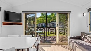 Une pièce moderne avec une table blanche, un canapé, un téléviseur fixé au mur et une porte-fenêtre coulissante donnant sur un espace extérieur avec vue sur une rue et des espaces verts.