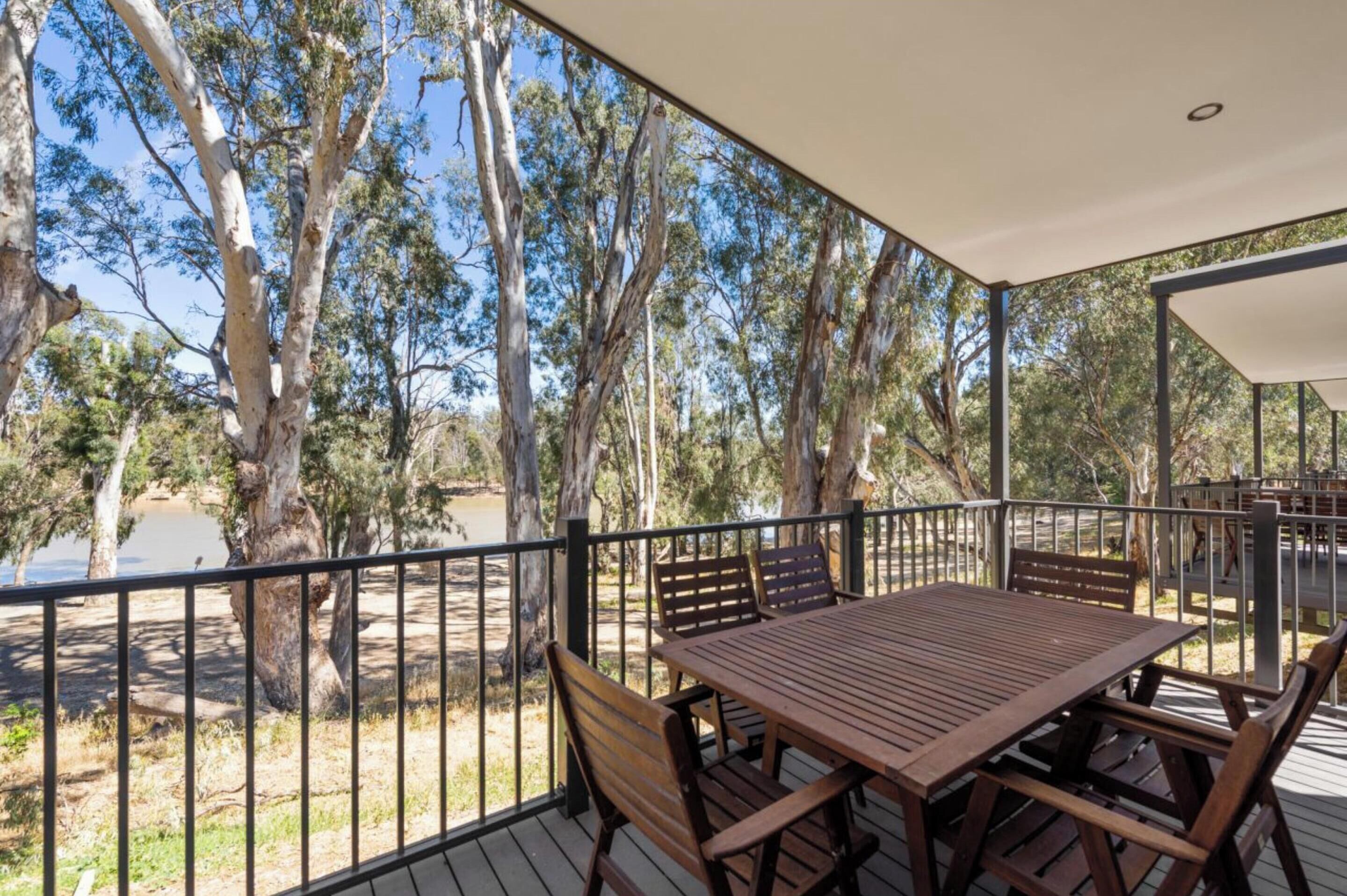 A wooden deck with a table and chairs, surrounded by a metal railing, overlooking a natural landscape with trees and a body of water.