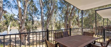 A wooden deck with a table and chairs, surrounded by a metal railing, overlooking a natural landscape with trees and a body of water.