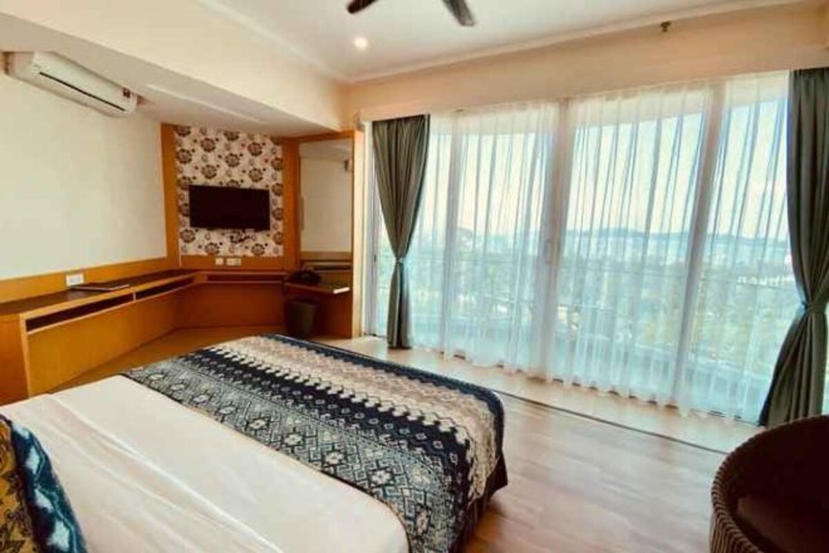 Ein Hotelzimmer mit einem großen Bett, einem an der Wand befestigten Fernseher, einem Schreibtisch und Blick auf die Stadt durch große Fenster.