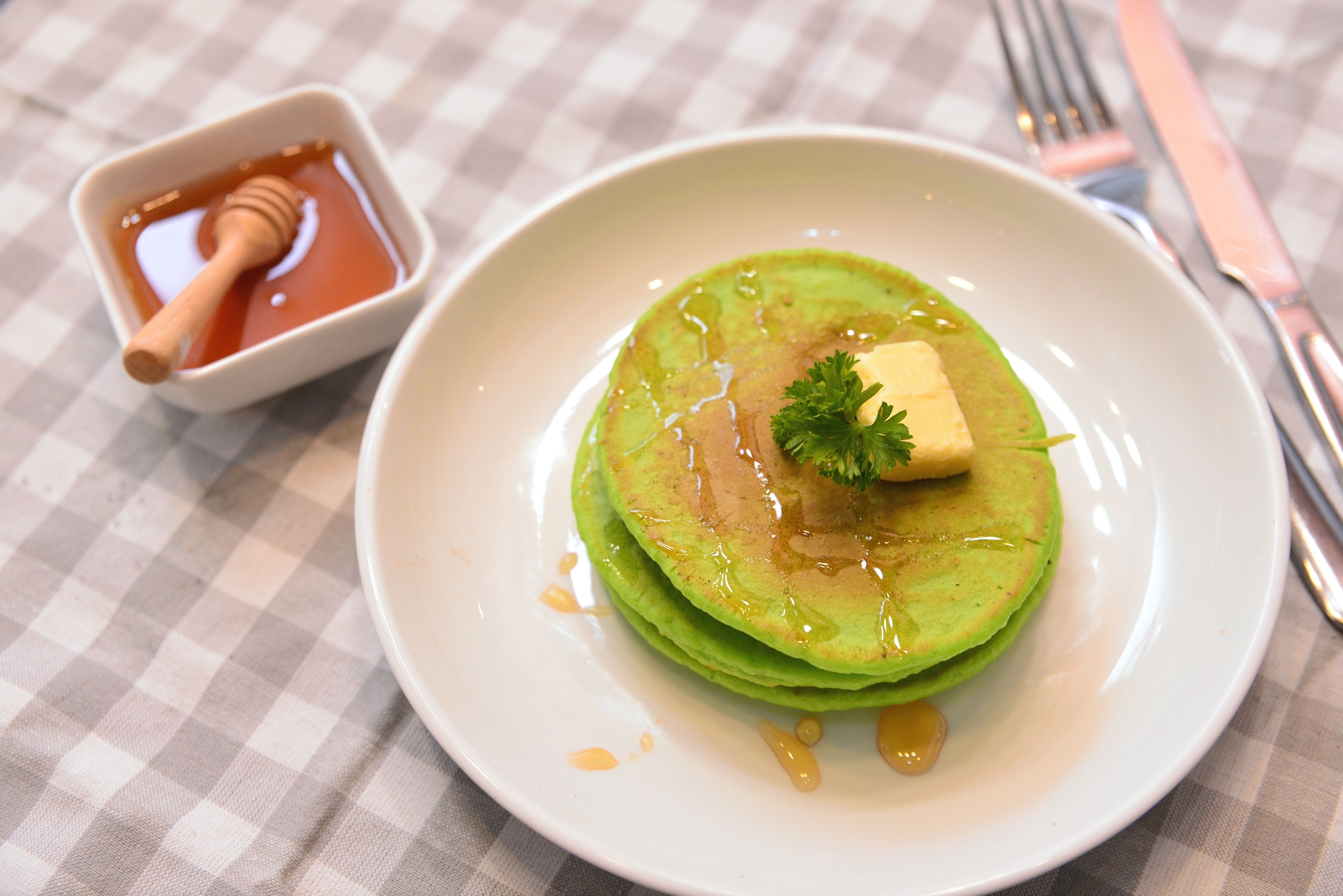 Une assiette de crêpes vertes, nappées de miel et garnies d’une noisette de beurre, servies sur une assiette blanche posée sur une nappe à carreaux.