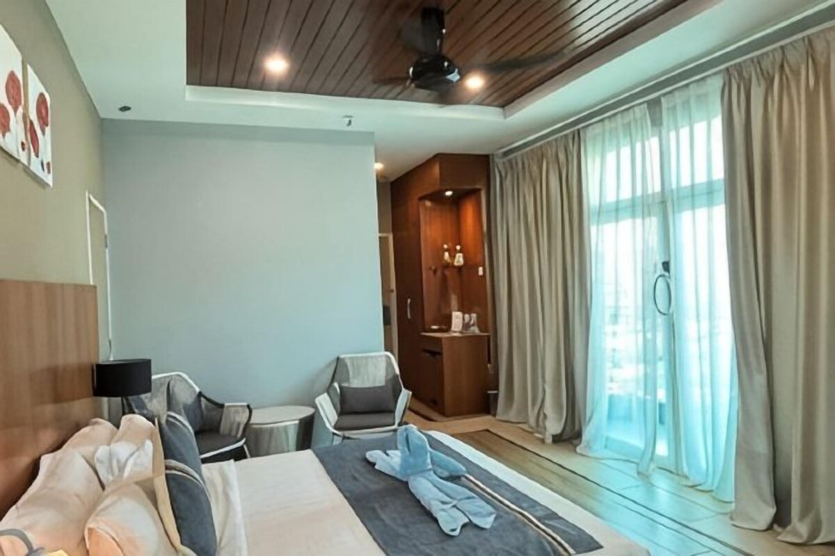 A hotel room with a bed, a seating area with chairs, a small table, a ceiling fan, and large windows with curtains.