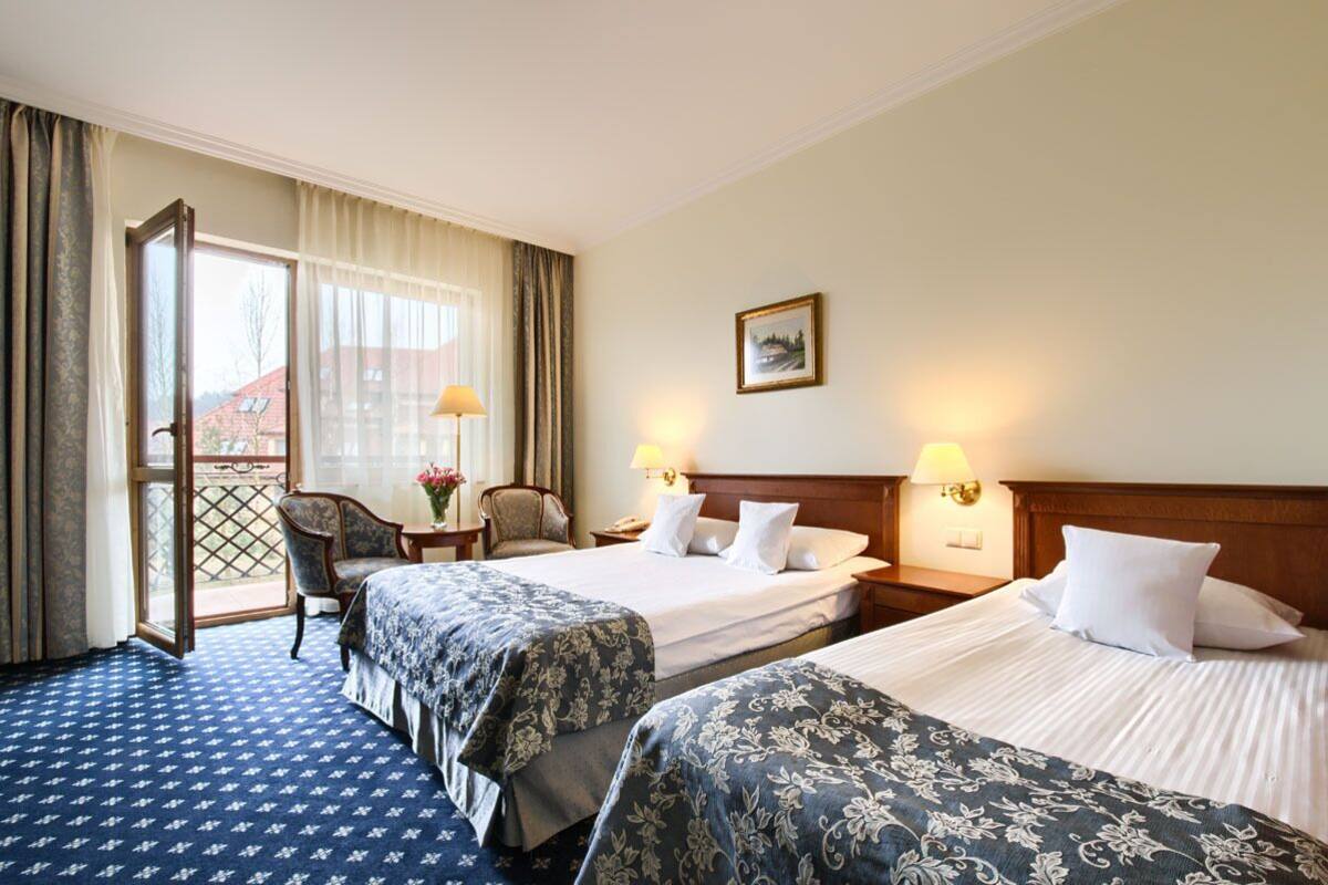 A hotel room with two beds, a chair, a table with a lamp, and a window with curtains.