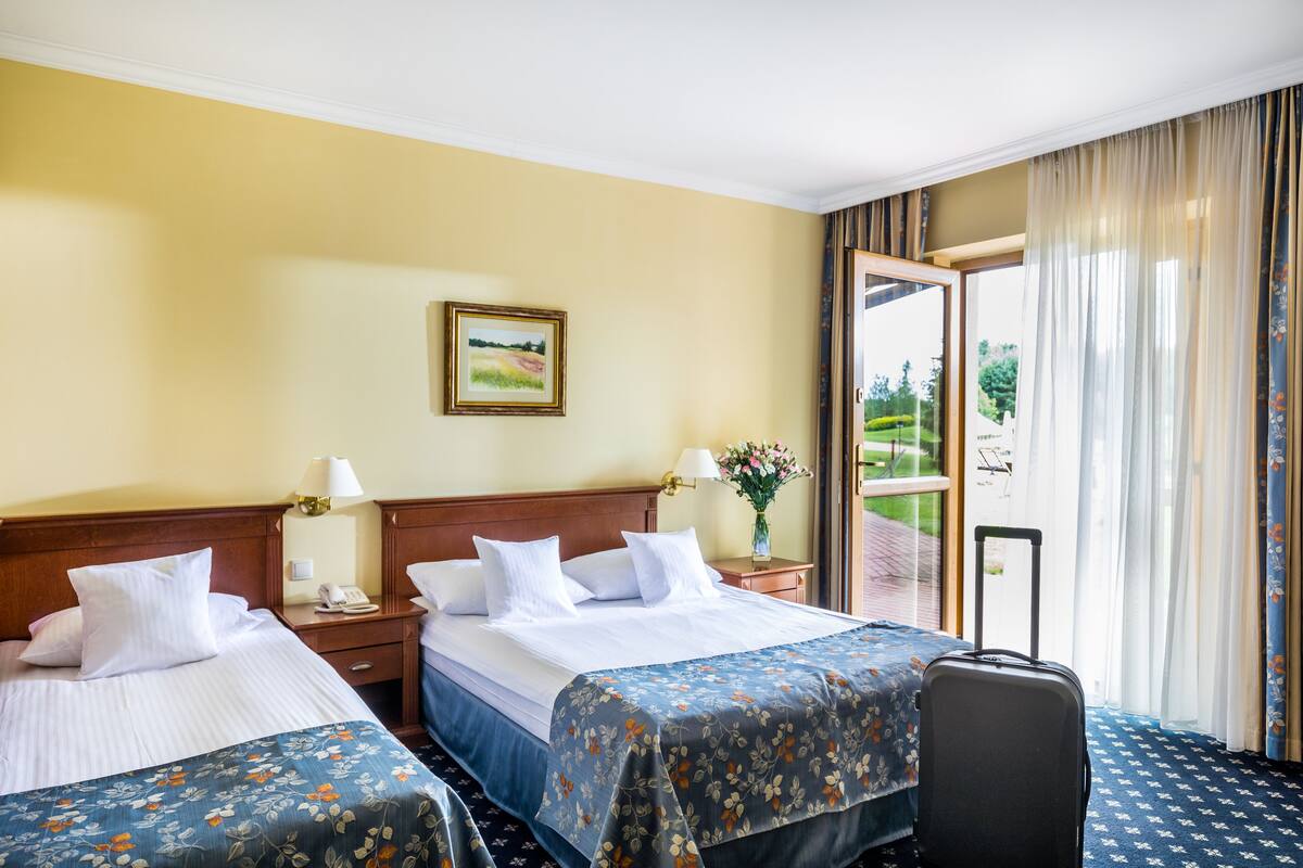 A hotel room with two beds, a wooden headboard, a nightstand with a lamp, a window with curtains, and a view of greenery outside.