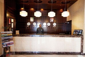 A hotel reception area with a marble counter, wooden paneling, multiple clocks, and a digital display.