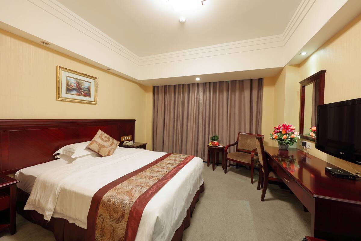 Une chambre d’hôtel avec un grand lit, un bureau avec une télévision, une chaise, une table de chevet et un tableau au mur.