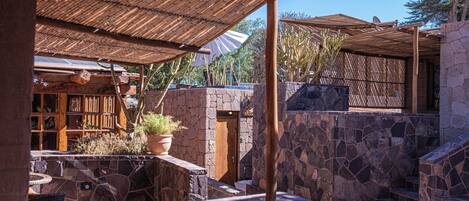 A stone-walled patio with a wooden pergola, a potted plant, and a building with wooden bookshelves.