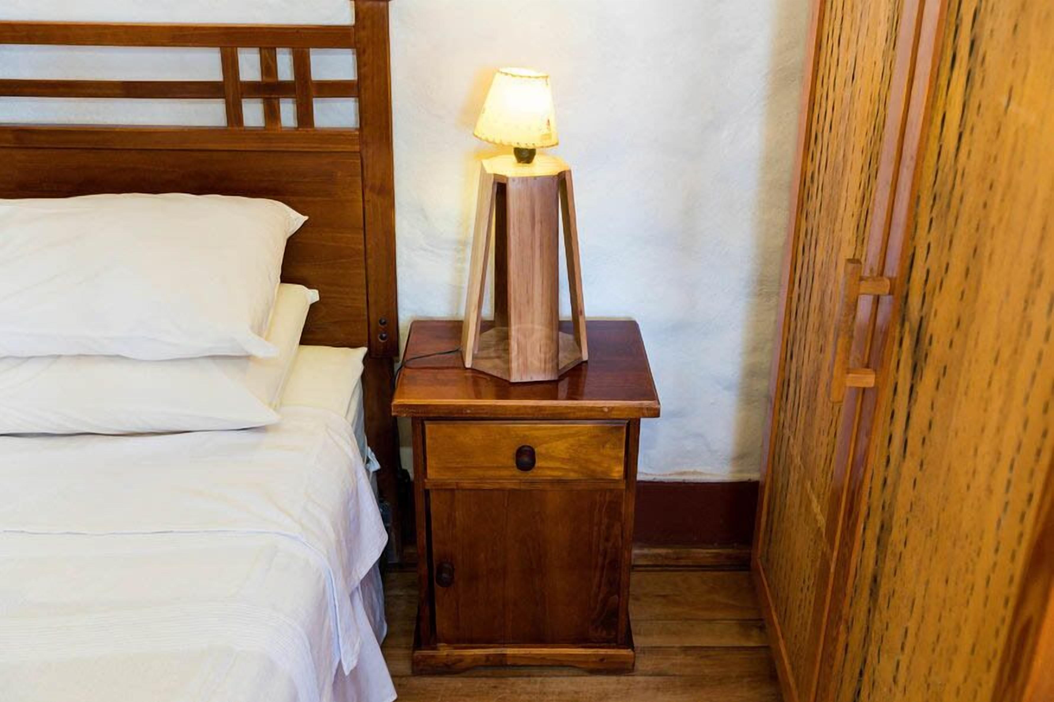 A wooden bedside table with a lamp, a bed with white linens, and a wooden wardrobe in a hotel room.