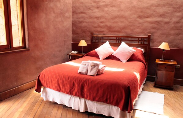 Una cama bien tendida con una colcha roja, dos almohadas y un par de toallas al pie. Hay dos mesitas de noche, con lámparas y un teléfono en una de ellas.
