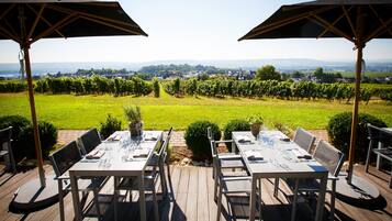 An outdoor dining area with tables and chairs, surrounded by a vineyard and a view of a town in the distance.