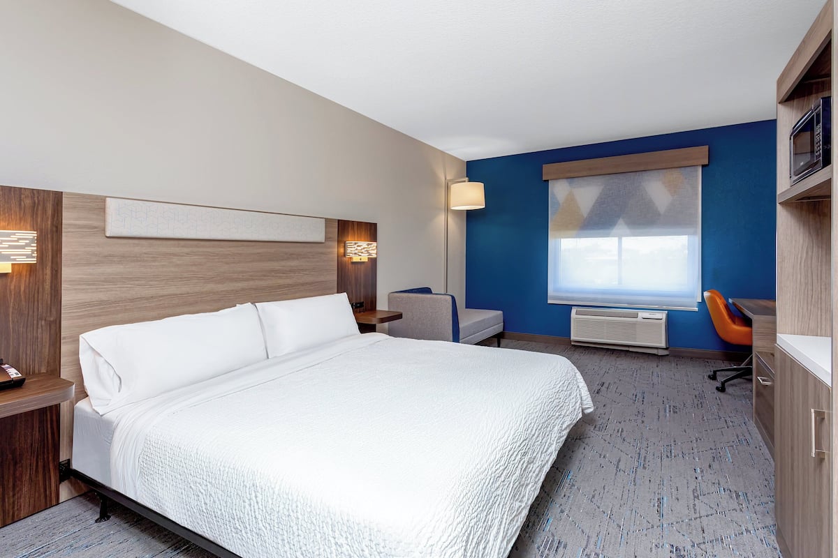 A hotel room with a large bed, a desk, a chair, a microwave, and a window with blinds.