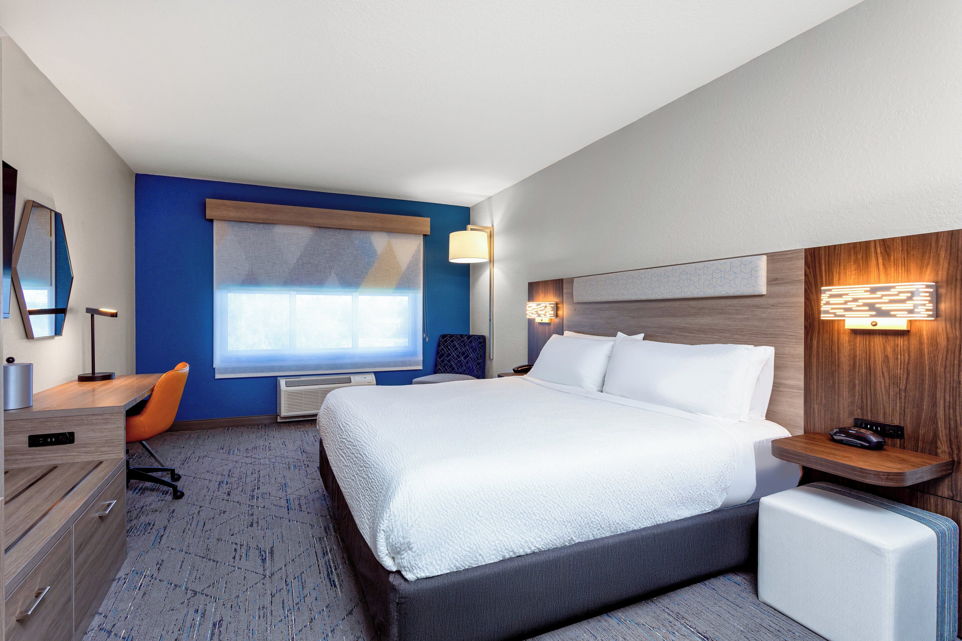 A modern hotel room with a large bed, a desk, and a chair. There is a window with a view of the outside.