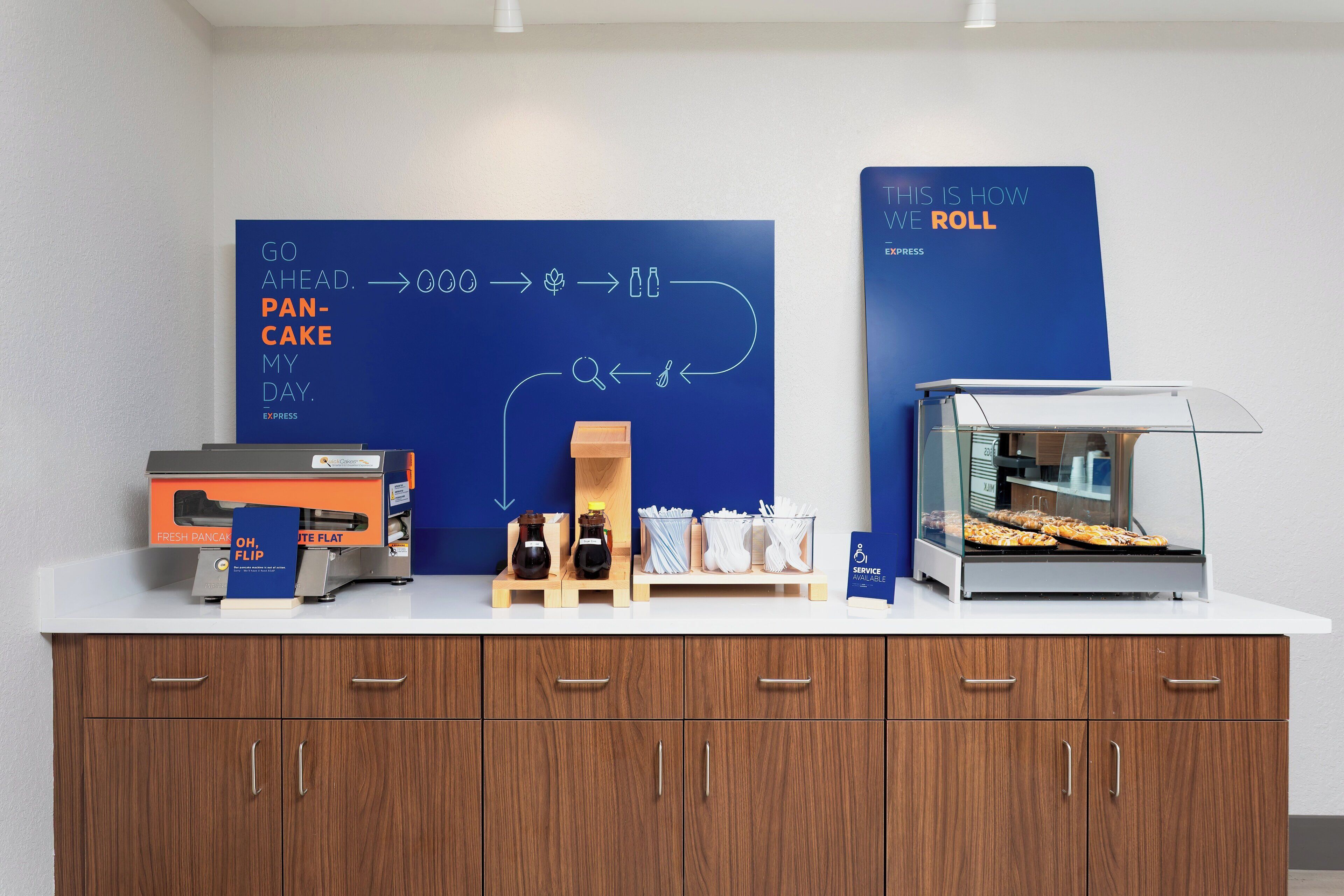 Una estación de desayuno con un cartel azul, una sandwichera y una vitrina con pastelería.