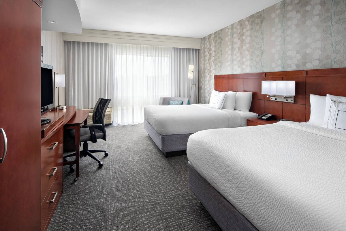 A hotel room with two beds, a desk with a computer, a chair, and a large window with curtains.