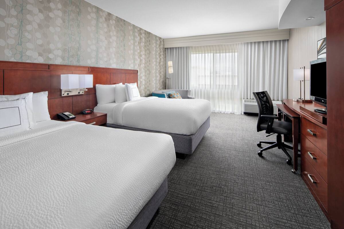 A hotel room with two beds, a desk, a chair, a TV, and a large window with curtains.